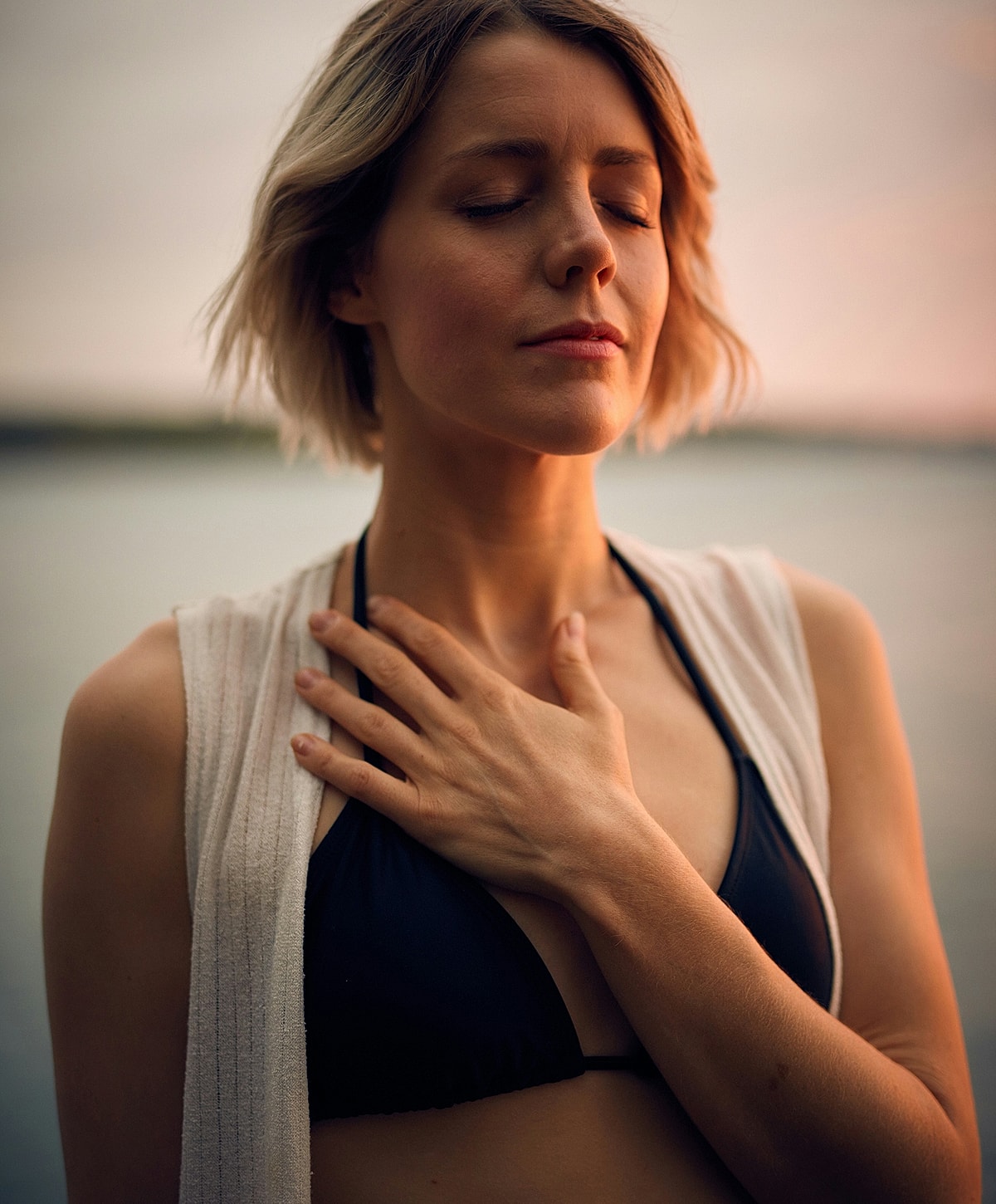 Woman closing eyes by water at sunset.